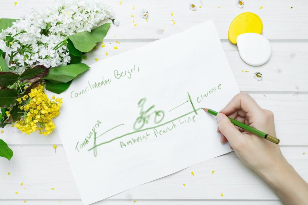 Tabletop with flowers and a piece of paper. A hand is holding a green pencil, drawing a bicyclist riding along a line representing the Amherst Peanut Line.
