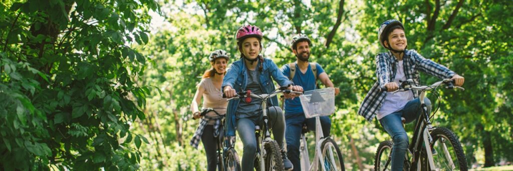 Two adults and two children riding bikes through sunlit foliage.